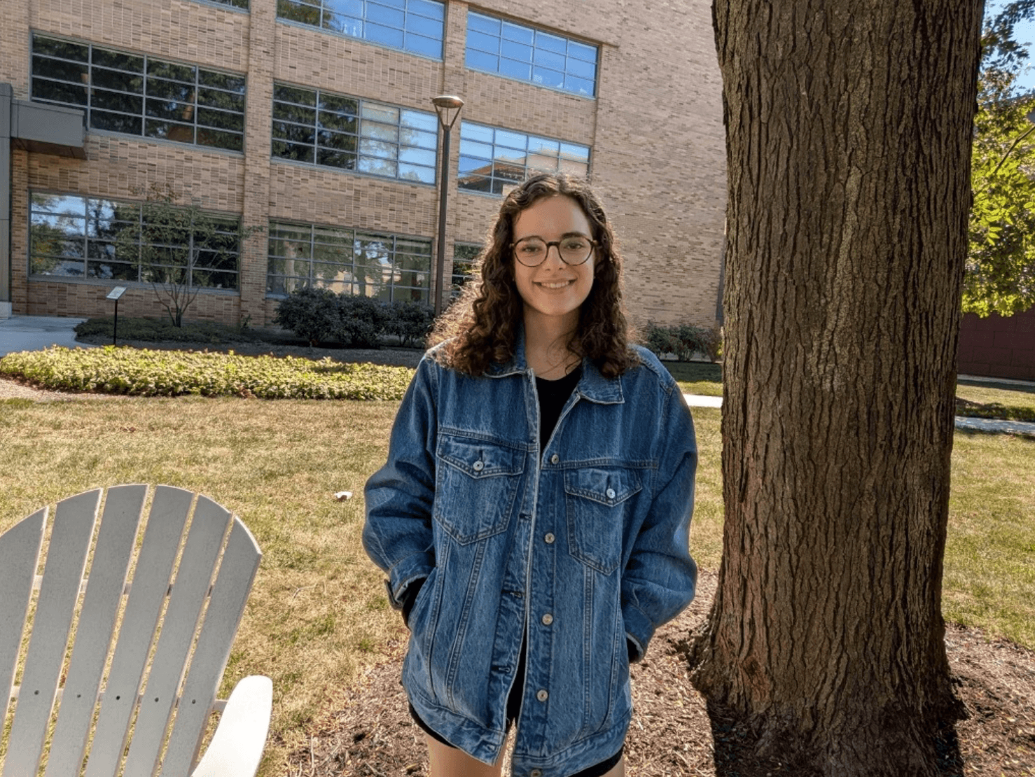 Mary wearing a blue jean jacket. She is standing between a white chair and a tree