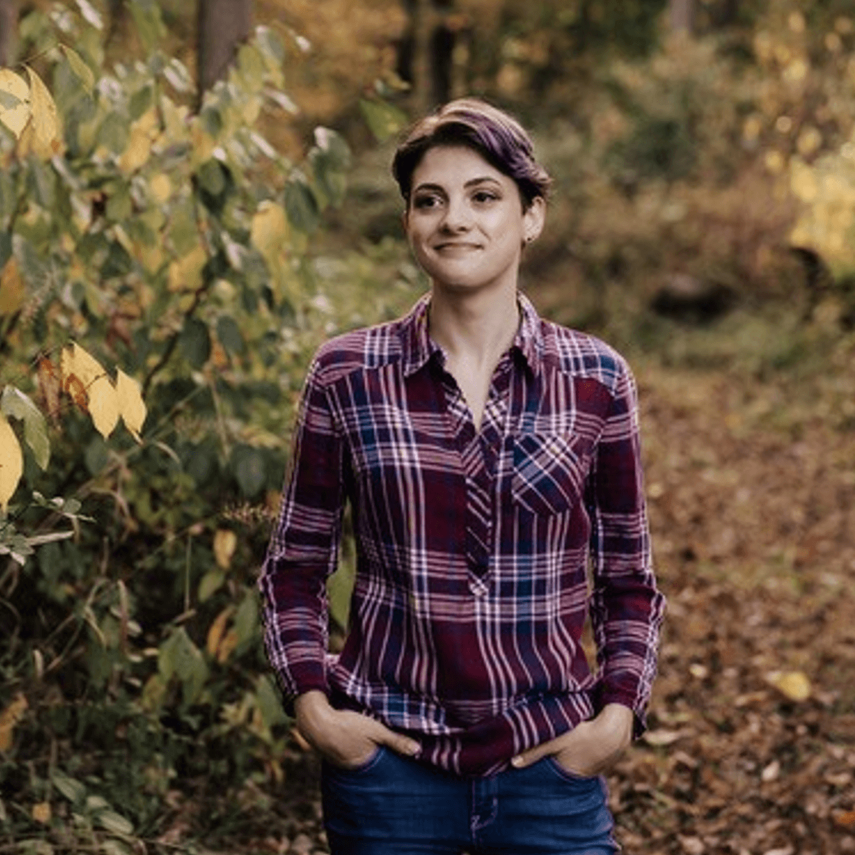 Sarah wearing a striped button-up. She is standing in the woods next to some leaves.