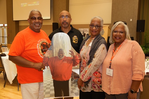 Four people pose with a large photo of a man