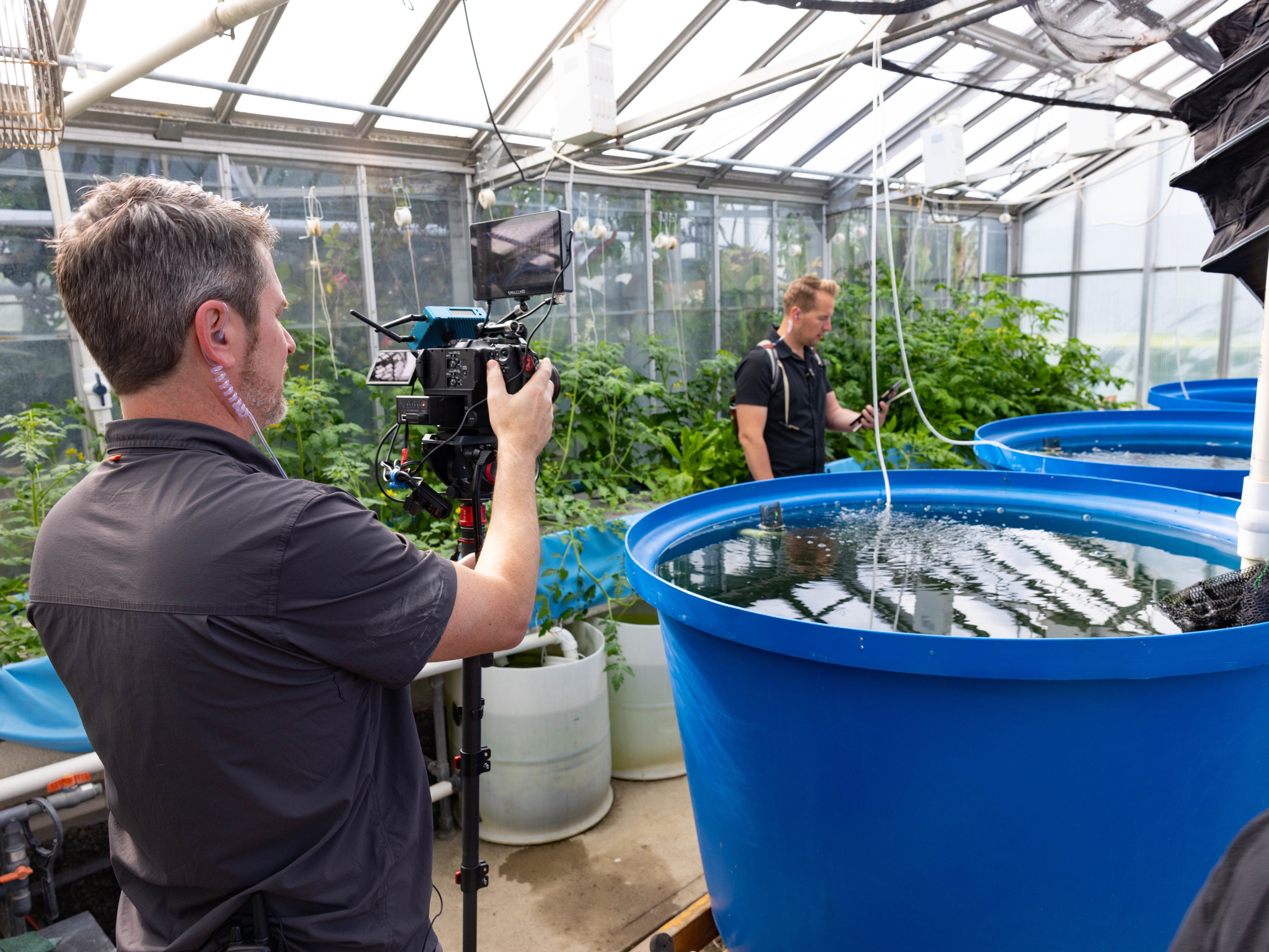 Two men with cameras record footage of a three-tank aquaponic system