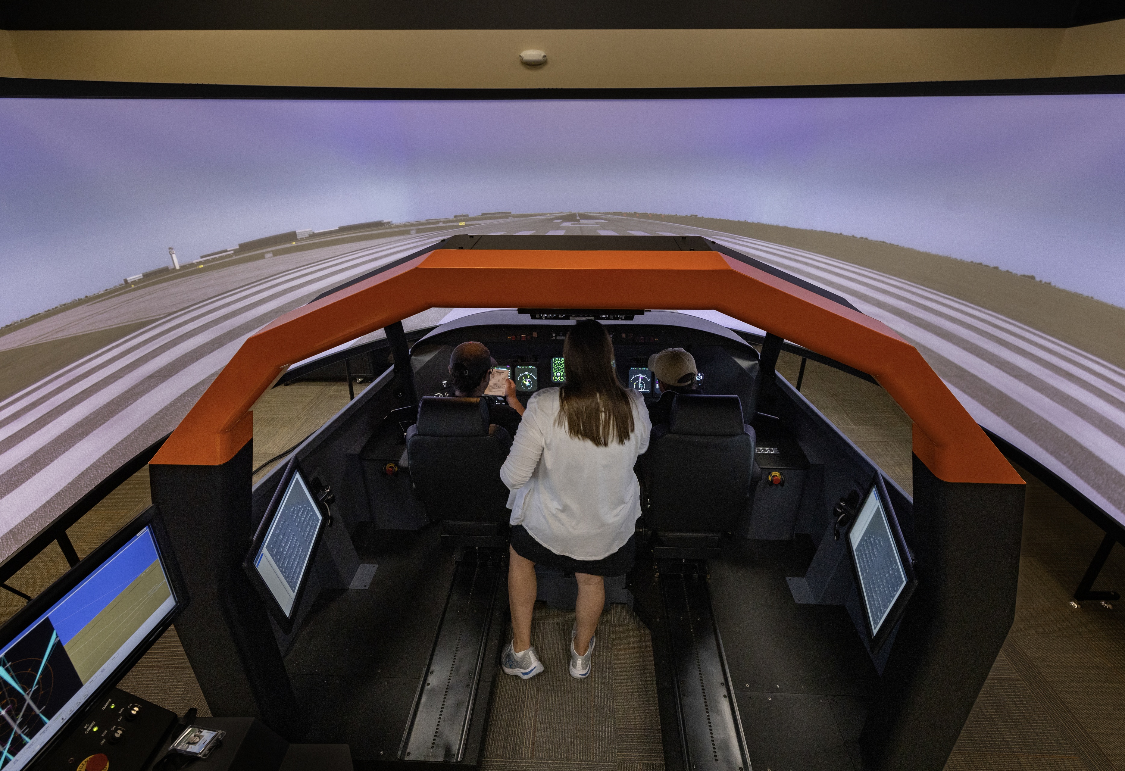 Two students and an instructor work inside flight simulator with a six-foot tall wrap around screen showing a virtual runway