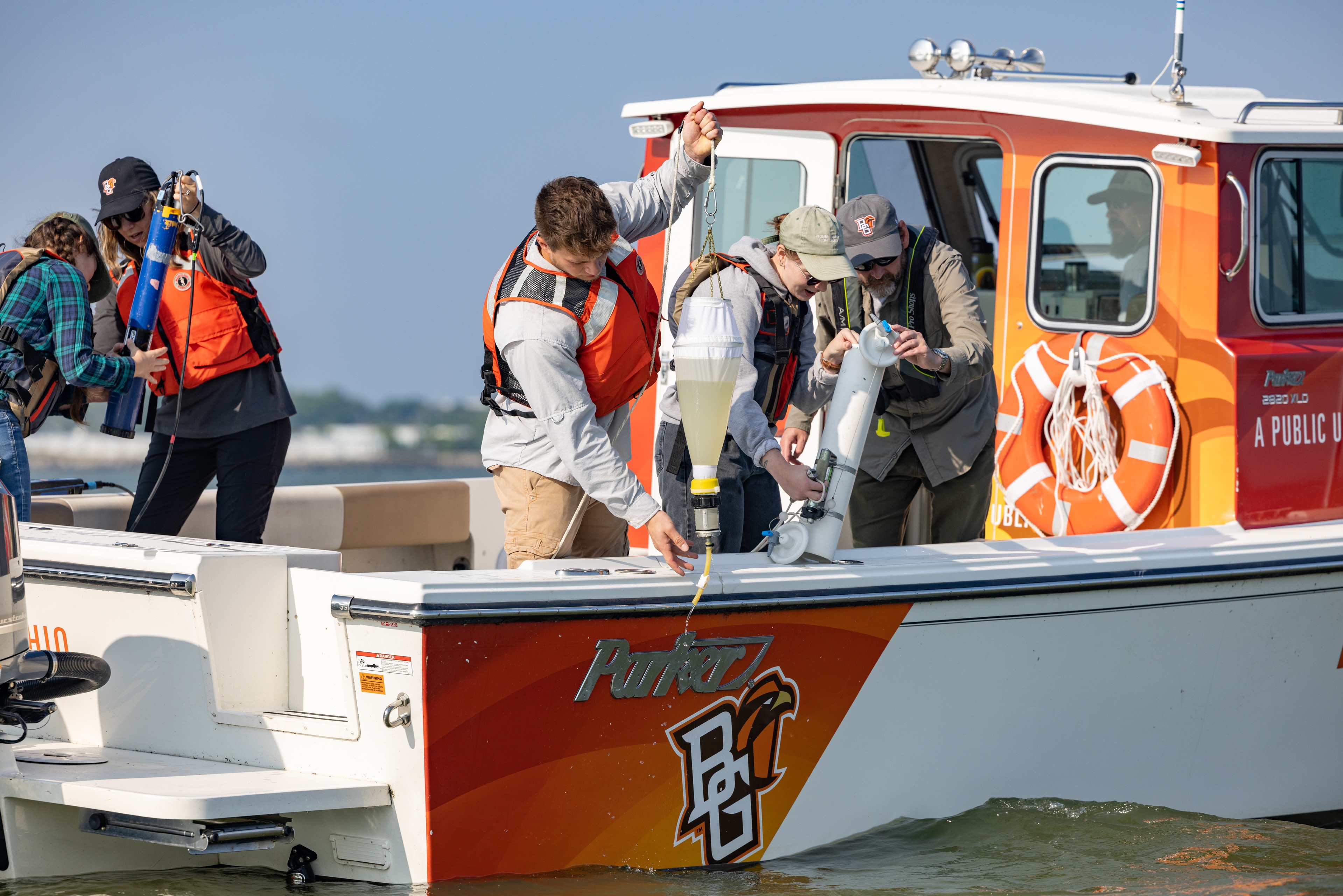 People wearing life jackets use equipment to pull water samples over side of boat