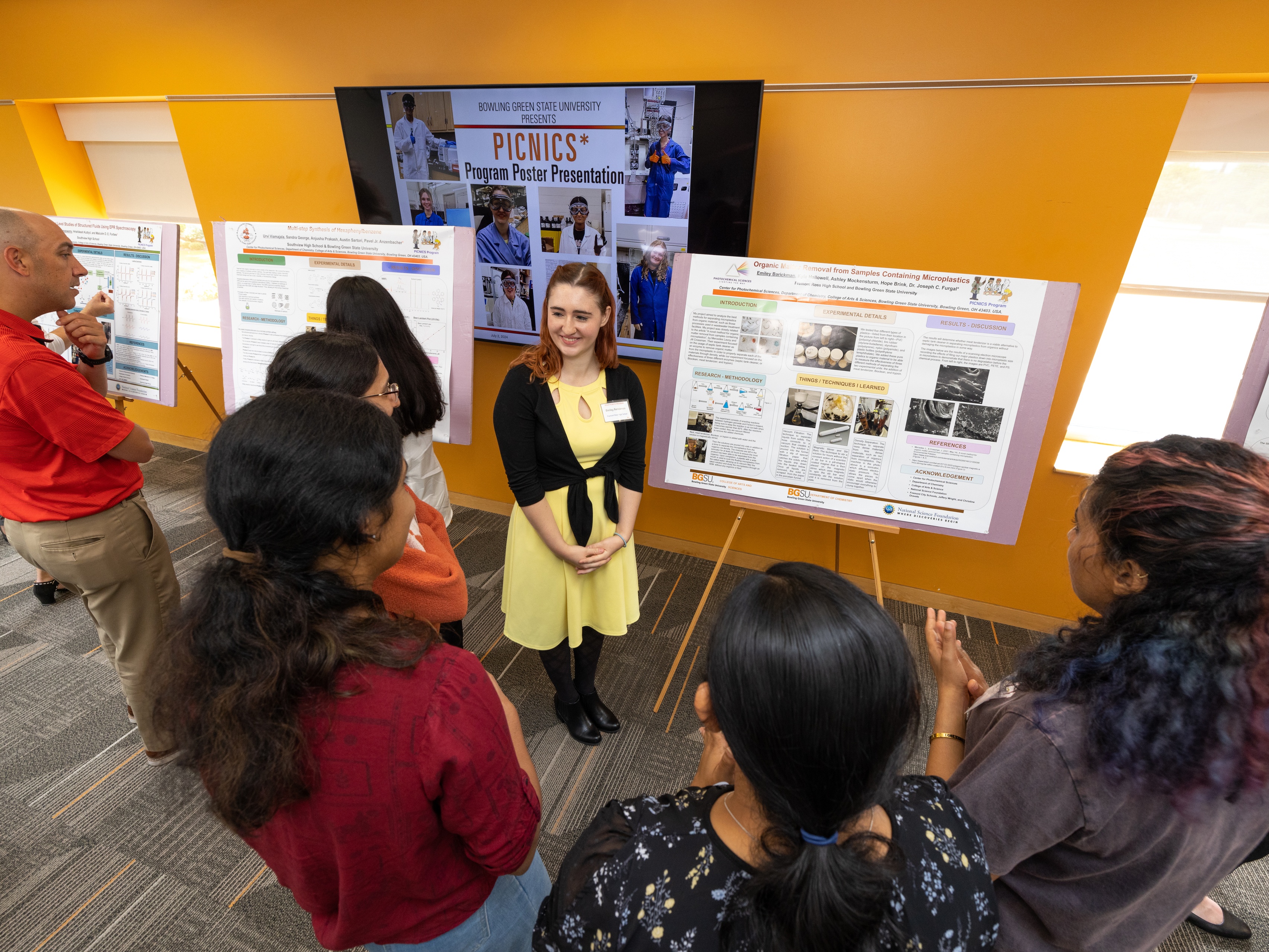 A high school student stands next to a board discussing her research with others
