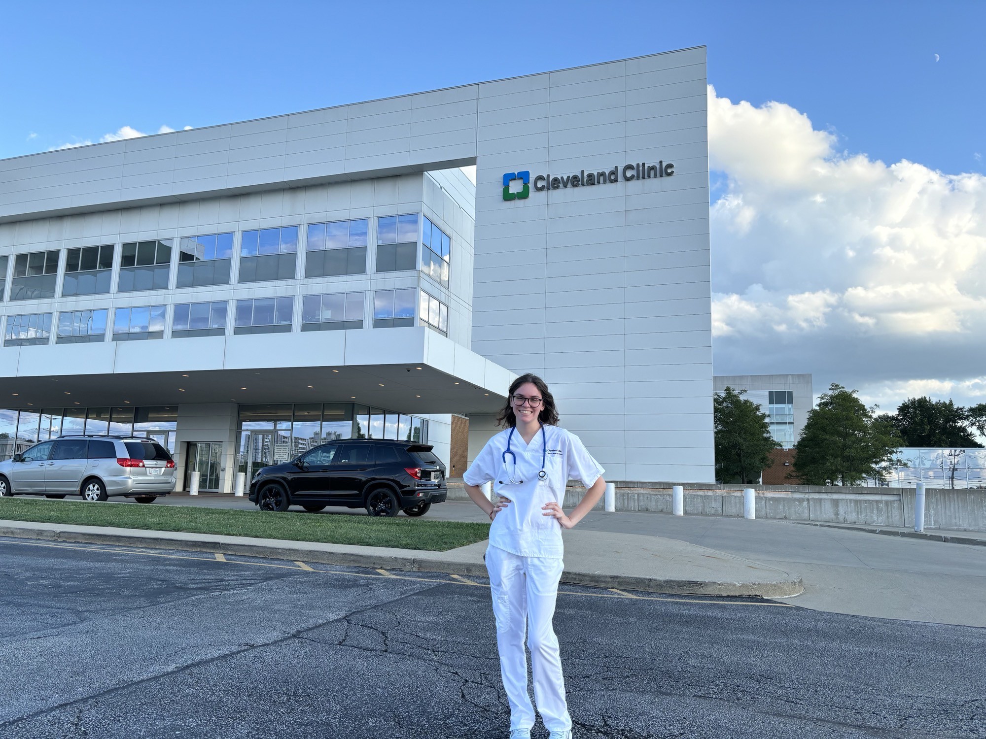 A BGSU nursing student wearing white scrubs stands in front of a Cleveland Clinic hospital.