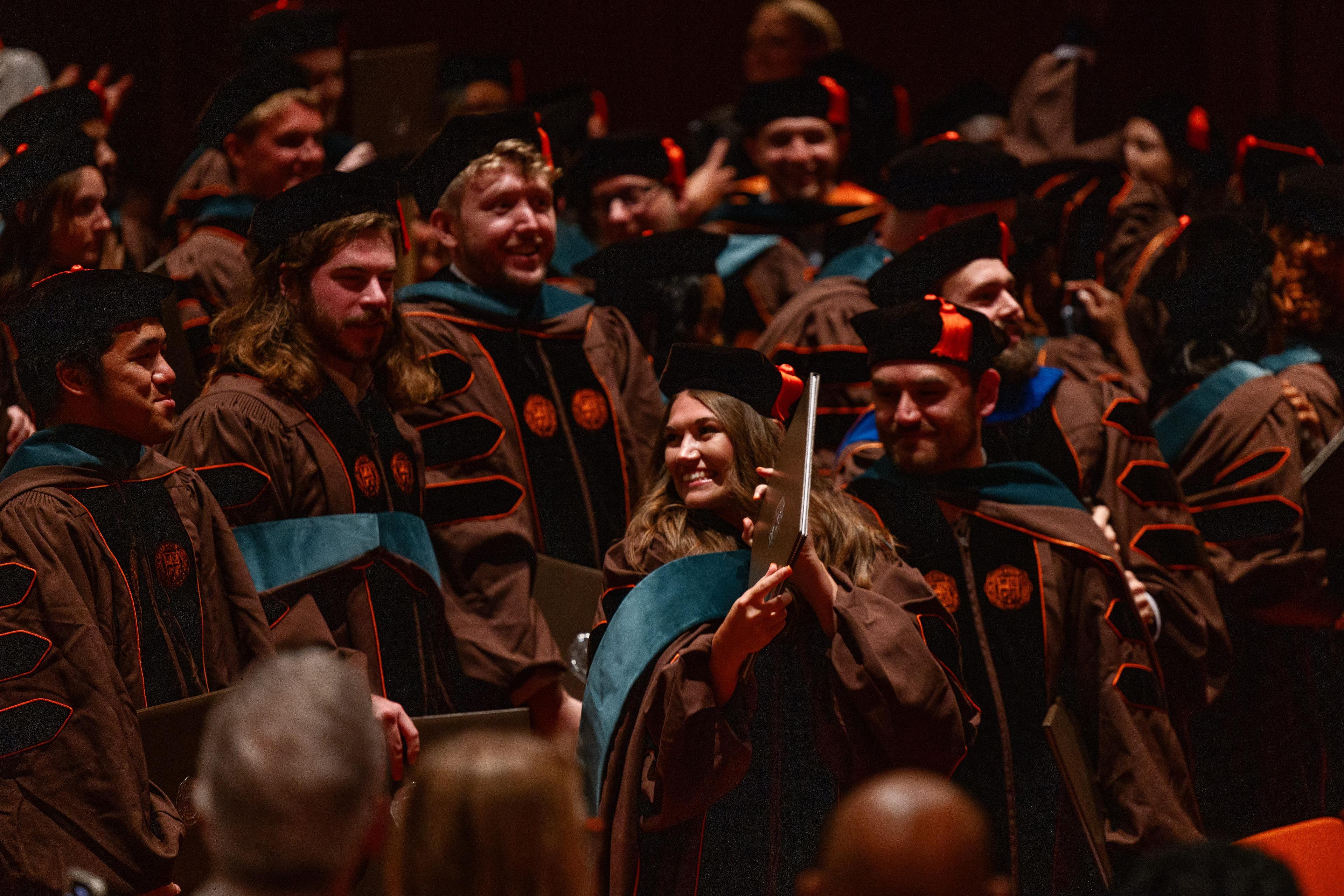 A graduate holds up her diploma and smiles while surrounded by other graduates.