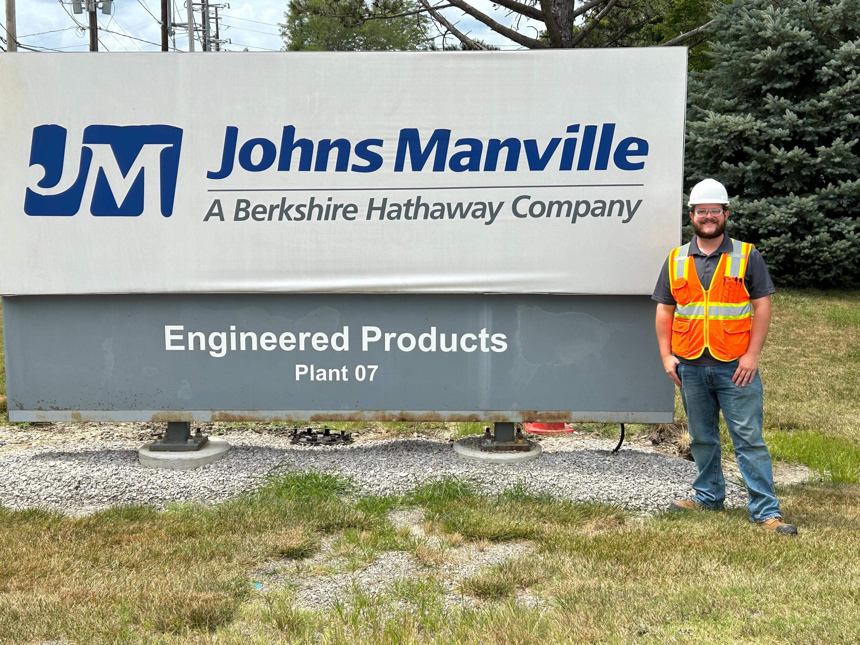 A BGSU student wearing a safety hat and vest stands next to a Johns Manville sign.