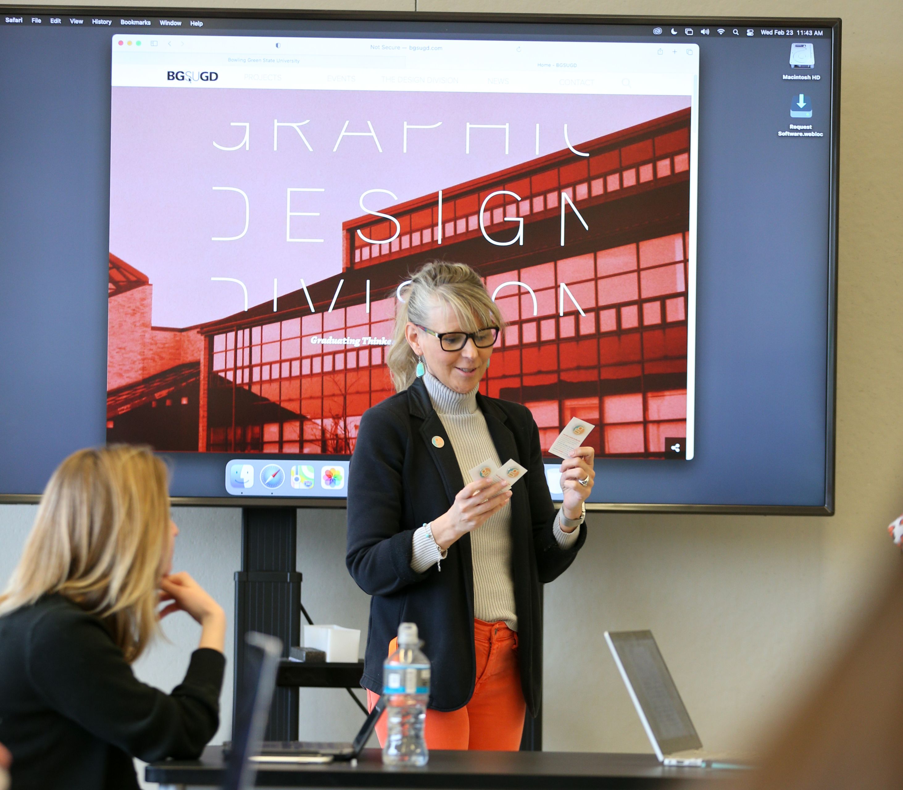 Professor stands in front of students with a TV screen behind her