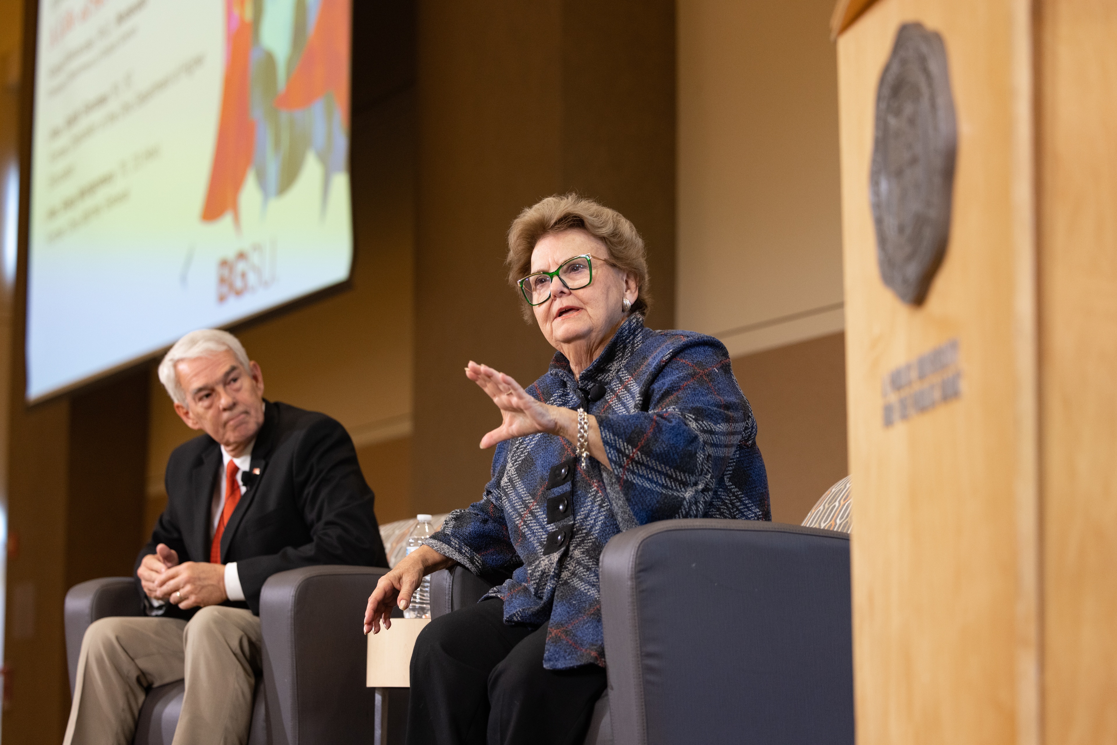 Two people sit in chairs on a stage.