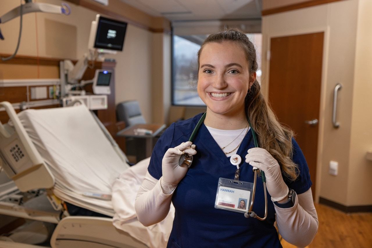 A nurse, wearing gloves and a stethoscope around her neck, smiles.