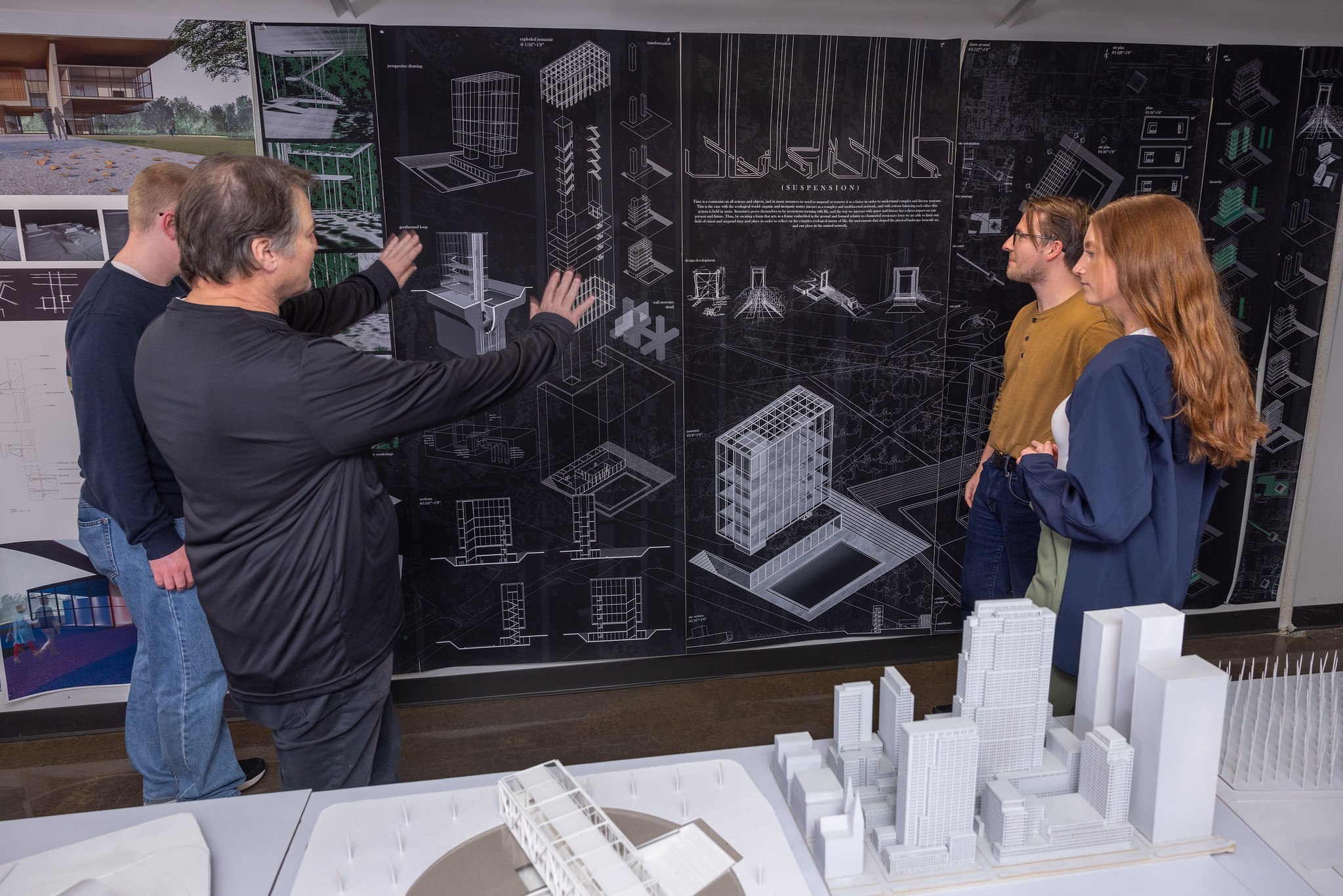 A professor gestures to a large architectural blueprint displayed on the wall with architectural models in the foreground as three students look on.